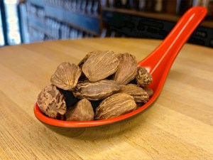 Cuchara llena de vainas de cardamomo negro, encima de una tabla de madera en la tienda de especias Sally Pepper en Madrid.
